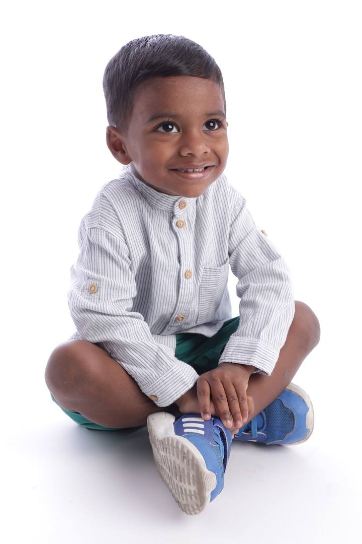 Murari seated cross‑legged on white background wearing striped shirt and blue trainers